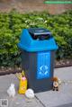 A blue trash can sitting on the side of a sidewalk.